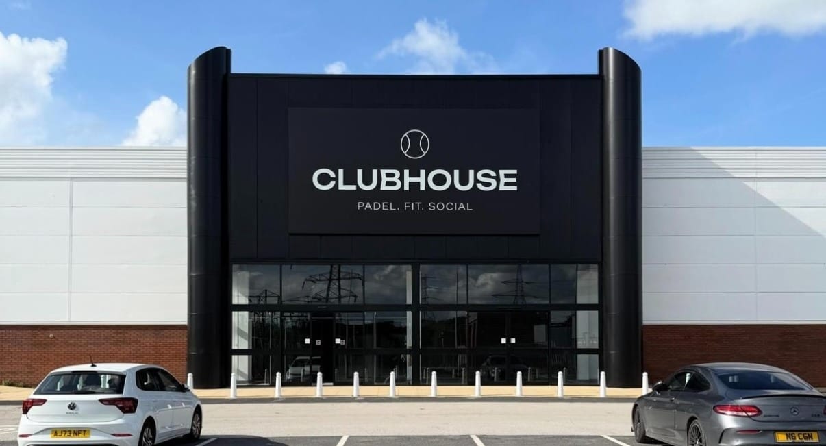 The entrance to Clubhouse Blackburn: a black two-storey entrance block with the Clubhouse sign and full-height glazing, set into the white cladding of the retail park unit, with cars parked in front
